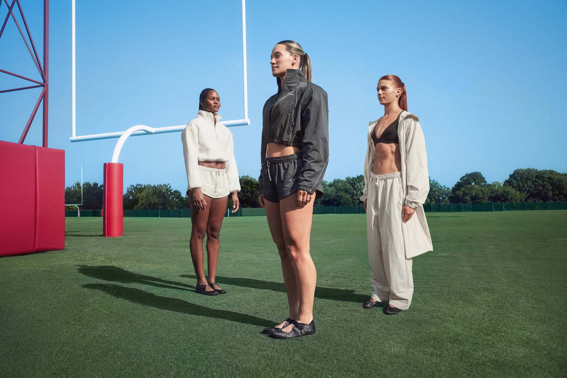 Three women wear NikeSKIMS outerwear on a football field.