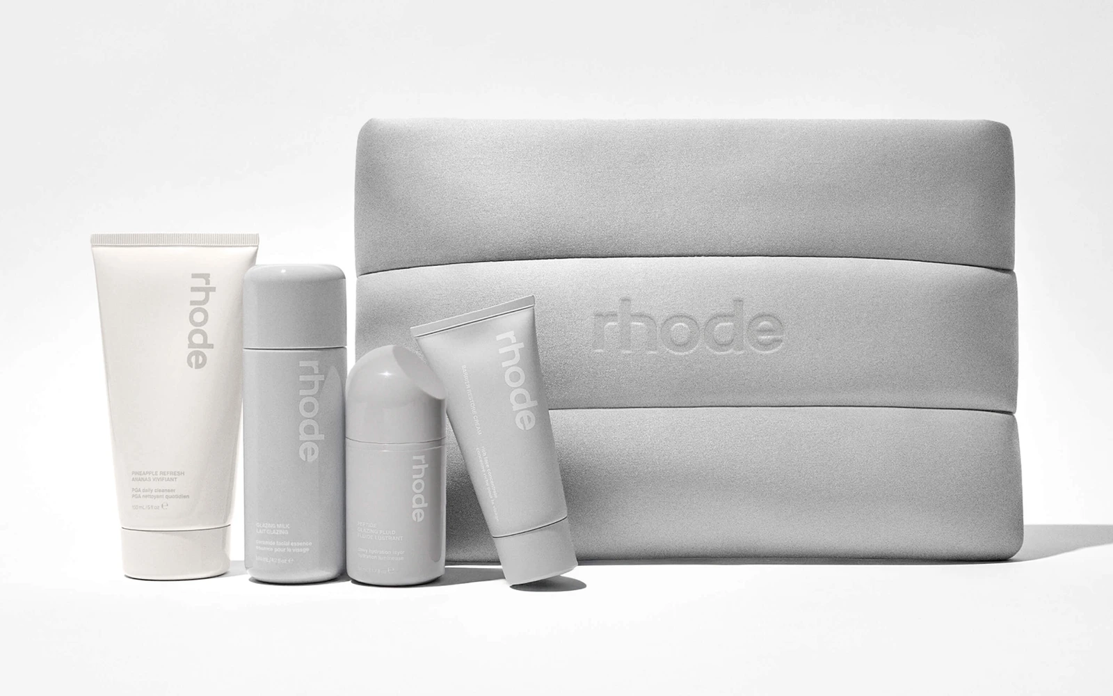 Rhode skincare bottles and tubes in front of the grey Rhode Kit pouch, photographed by the brand.