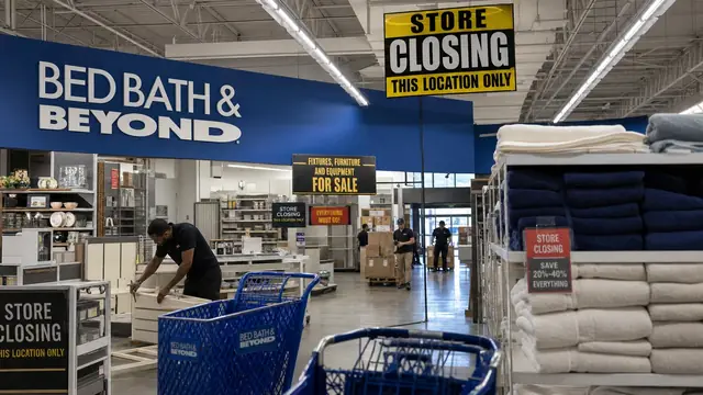 Bed Bath & Beyond store-closing liquidation scene with blue storefront sign, pallets, and staff handling inventory.